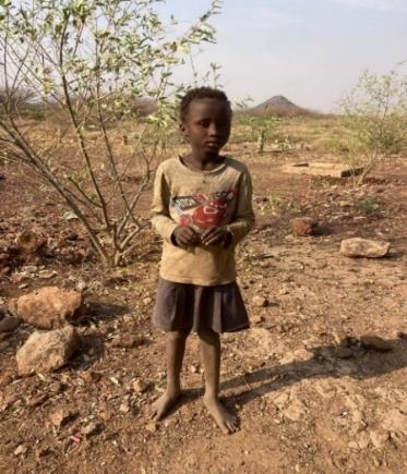 A young child stands barefoot among dry thorn bushes near Kakuma
