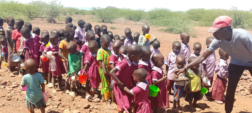 Village children in pink school uniforms queue with bowls and cups for a lunchtime meal